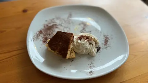 A white plate of tempting-looking tiramisu, dusted with cocoa on a wood-topped table