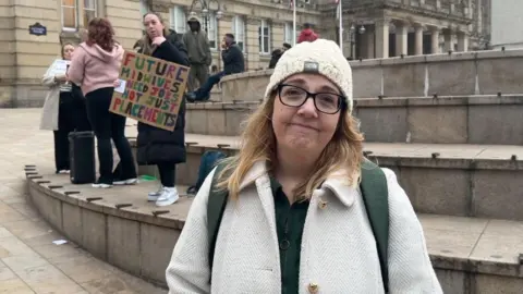 A woman with a hat, shoulder length blonde hair and glasses is looking at the camera and smiling. She is wearing a white coat and is standing in front of some stone circular steps, next to other campaigning students holding signs