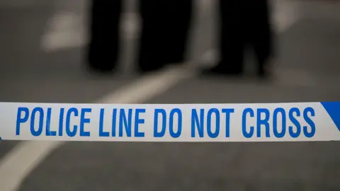 Police tape goes across the road and says in blue writing 'POLICE LINE DO NOT CROSS'. There are several police officers standing on the road in the middle.