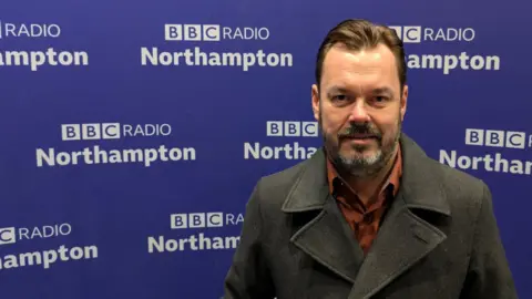 Joe Ashmenall/BBC A man in a burnt orange shirt and a charcoal pea coat stands in front of some purple BBC Radio Northampton branding. 