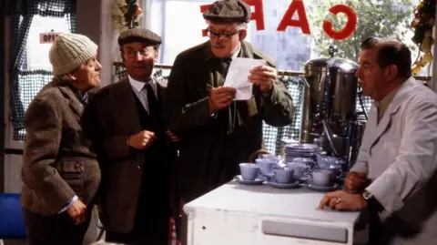 Group of elderly men look at each other as one reads a note to a quizzical looking cafe owner