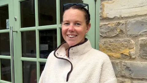 Tanya Jones is standing by a green door. She is wearing a white fleece and is smiling at the camera.
