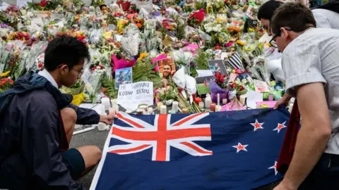 Getty Images Mourners lay out a New Zealand flag next to a bed of floral tributes for Christchurch shooting victims in 2019