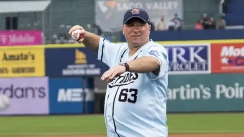John Lewis threw the ceremonial first pitch in front of thousands of fans. He is wearing a pale blue a baseball top with dark letter on it and dark coloured shorts and a dark baseball cap. He is standing on the pitch with sports stands behind him.