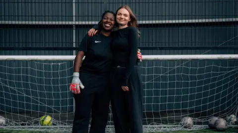 Morgane Bigault / Comic Relief A black woman and a white woman embrace in a football goal with several balls behind them