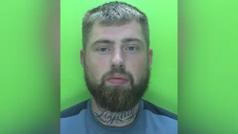 A custody photo of Luke Williamson, a white man with a large neck tattoo and a short beard with short blonde hair.