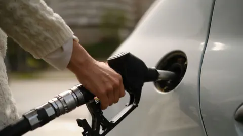 A woman using a gas pump to refuel vehicle during energy crisis. She has a silver car, and is wearing a white sweater.