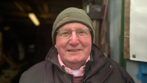 Dave is wearing a dark green beanie hat that has orange lettering reading "Bartley Park". He wears glasses, and a brown coat with a wide collar over a pink shirt. He is looking at the camera and smiling, and a small shipping container full of tools like pitchforks is in the background, but out of focus.