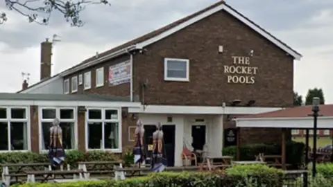 Google The pub has a garden at the front with tables and chairs outside and with its sign on a wall at the entrance to the pub.