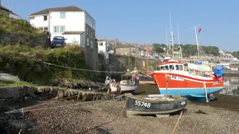 A boat tied up on sand which is made of pebbles, the boat is blue and red and next to it is a smaller boat also tied up on sand that is black and says SS748 on it