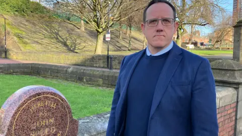 Man in blue suit stands beside a stone memorial to Sgt John Speed