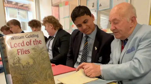 BBC Mick (right) sits with current pupils of the school. Mick is wearing a light blue suit and a tie, and is looking through an old text book with a pupil who is smiling. Other pupils, all wearing black blazers, blue shirts and blue striped ties, are in the background. In the foreground there is a copy of Lord of the Flies, with a jungle scene on the cover. 