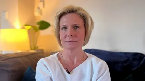 BBC A woman with blonde hair, wearing a white top, sitting on a grey sofa, with blue cushions, with a lamp and two house plants in the background.