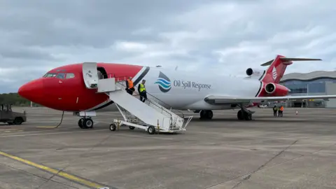 Red and white Boeing 727 aeroplane branded 'Oil Spill Response' at Doncaster Sheffield Airport, with two men in hi-vis jackets descending the steps.