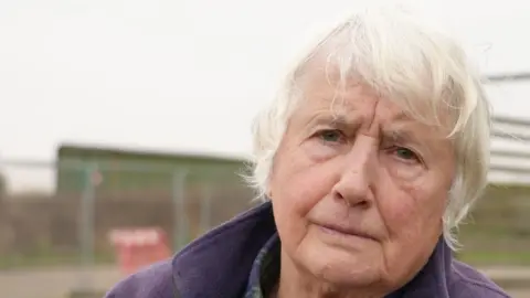 Bryony Nierop-Reading, a woman with short grey hair. She is wearing a blue and green checked shirt with a cream jumper. She is also wearing a purple fleece and is looking into the camera.