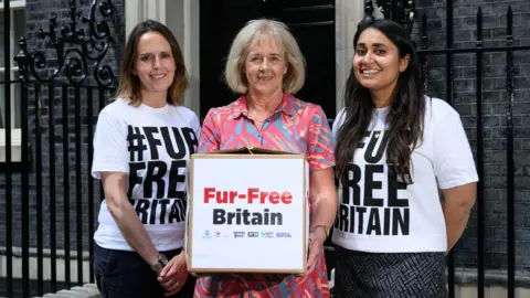 Doug Peters/Humane World for Animals Ruth Jones stands beside two women wearing white shirts that say #FurFreeBritain. Ms Jones is holding a cardboard box with the same words on the outside. She is smiling and is wearing a pink and blue patterned shirt. She has shoulder-length blond hair. The woman to her left has shoulder-length brown hair and is smiling. The woman to Ruth's right has long dark-brown hair and is also smiling. The trio are stood outside Number 10