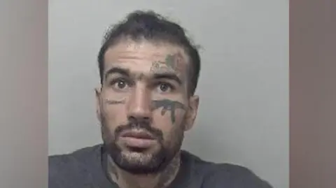 Metropolitan Police Custody photo of Fayaz Khan, who has short dark hair, a beard and moustache. He has tattoos on his face, including one of an AK-47 gun beneath his left eye. He is wearing a grey jumper.