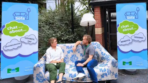 A sofa with a blue and white cover. A women and a man are sitting on the sofa and talking. There are two signs explaining what The Sofa initiative is 