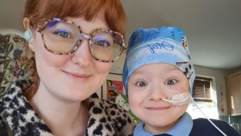 Amy Dixon A picture of Amy (left) wearing glasses and smiling and Freya (right) also smiling, wearing her school uniform and a bandana with a tube in her nose.