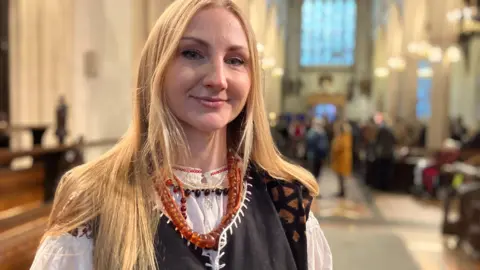 Jon Wright/BBC Alina Dolgopolova stands inside a church, smiling at the camera. she is wearing a dark tunic and a white shirt.