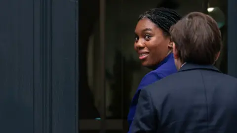 PA Kemi Badenoch walks towards the entrance of a building, turns and looks over her shoulder at the camera