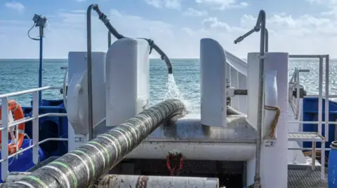 A black and green striped thick interconnector cable going off the back of a ship into the sea. There is a hose on top of the cable spraying water on it. 
