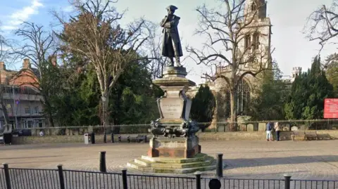Google Public square with black railings surrounding a stone monument with a figure on the top wearing a tricorn hat and long dark coat. He is standing on a plinth on which his name, John Howard, is inscribed. There are three steps at the base of the statue. There is a church behind the statue, to the right, and a tree to the left.