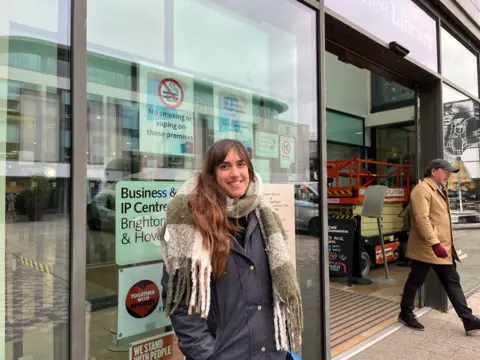 George Carden/BBC Becky Netley stood by the entrance to the glass walled Jubilee Library in Brighton. A man is walking out of the library. Becky is wearing a navy blue jacket and large green and white chequered scarf. 