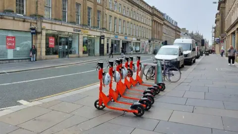 Graham Robson/Geograph Six orange e-scooters are lined up on the pavement in Grainger Street, Newcastle. Shops, including a sports shop, are on the other side of the road. 