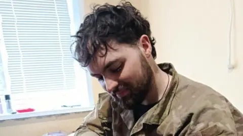 Natasha (Ayrton's mother) A bearded person wearing a camouflage outfit is sitting indoors.