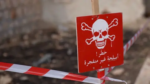 Red and white tape cordoning off a field, close-up of a red sign with a white skull. Sign reads: "danger, unexploded weapon",