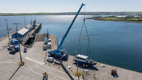 Colin Keldie/EMEC A small ferry being lifted by a crane from the deck towards a harbour with shimmering blue water