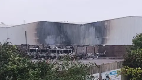 Spotted in Thornbury BS35 Burnt up lorries parked outside a warehouse. The side of the building is black from the fire and the lorries are badly damaged. 