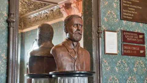 MANX SCENES A bronze bust of Frank Matcham, which has short hair, a moustache and beard and is dressed in a jacket and tie. There is a large wooden framed mirror behind the bust which is showing a reflection of its back. The wall is covered in highly decorated green coloured wallpaper and there are wooden signs pointing to the seating areas on the right.
