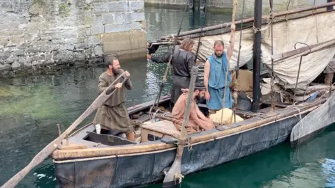 Brioc, a leather boat, is in calm waters in a harbour, with high stone walls behind. On board are several crew members in 10th Century clothing, with ropes and a sail to the right. On the left at the back of the boat the captain is holding a large wooden rudder.