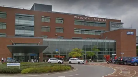 Google The main entrance to Whiston Hospital can be seen in this image from Google. There are a number of cars parked outside and there is a large glass panelled area at the front.