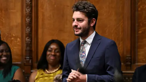 UK Parliament Jack Abbott standing and asking a question in the wood-panelled House of Commons. He has dark hair and a short bear. He is wearing a dark blue suit, white shirt and flowery tie. His hands are clasped in front of him and he appears to be speaking. People behind him are smiling.