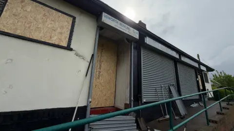 BBC/Jim Scott The club house, which has since been burnt out, is seen boarded up with wooden panels. Parts of metal shutters lay outside after being ripped down by firefighters who were responding to the incident.