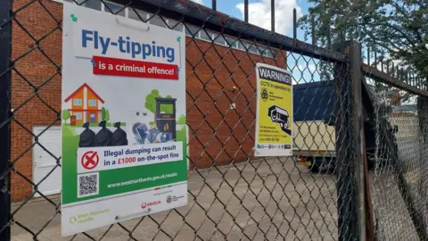 A close up shot of a wire fence, with a white poster strapped on. It says "Fly-tipping is a criminal offence. Illegal dumping can result in a £1000 on-the-spot fine. www.westnorthants.gov.uk/waste". There is another yellow poster warning people that CCTV is in operation.