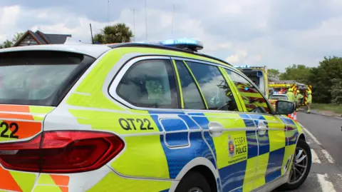 Police car in foreground with officers in background and more police cars on a road. A fire engine can also be seen.