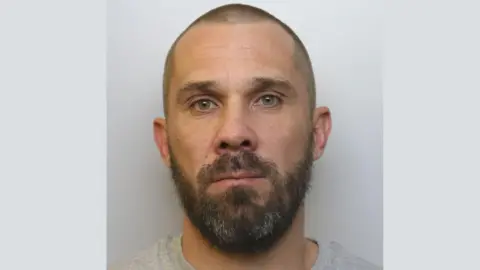 Avon and Somerset Police A police mugshot of David Garland. He is wearing a grey t-shirt and standing against a grey background. He has green eyes, a full dark beard and a shaven head. He is looking into the camera with a blank expression.