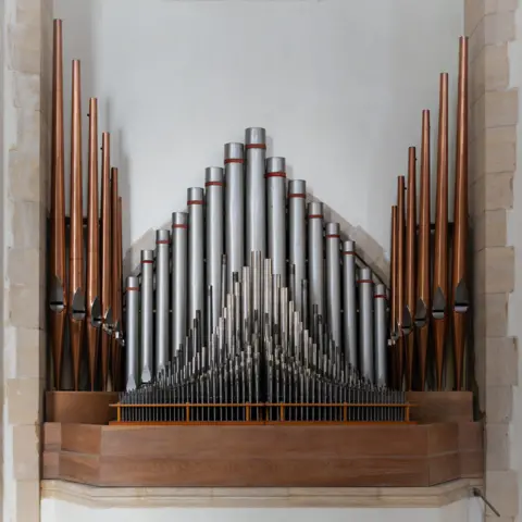 Marie Davey Dozens of organ pipes in varying sizes, silver and bronze in colour. The pipes are set into a white wall with stone brick trim.