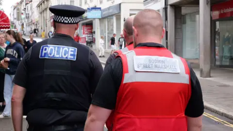 Three men from behind, one wearing a police uniform and two wearing red Bodmin street marshal vests patrolling a town street with people in front of them.
