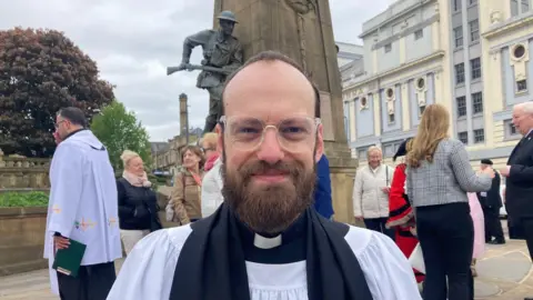 A man in glasses wearing the black and white robes of a Catholic minister standing in front of the Bradford War memorial