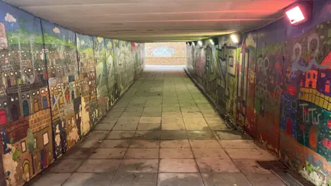 Toby Friedner/BBC A pedestrian underpass with tiled flooring and low ceilings, leading toward a brighter exit at the far end. Both walls are covered with colourful but worn murals depicting buildings and abstract designs, partially obscured by graffiti and peeling paint. Overhead lights cast a slightly uneven glow, with some fixtures appearing dim or discoloured. 