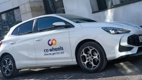 Co Wheels The picture shows a white Co Wheels car parked on a street in front of a modern building. The car is a small hatchback with the Co Wheels logo on the side, along with the words “Hire me, get the app”. The car has five doors, silver alloy wheels and a clean, polished exterior. 