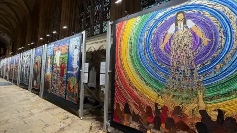 Textile thread displays held up by square panels in a minster building with stone flooring and stained glass windows that are dark. The display on the far right is colourful and depicts swirling colour with people stood below the depiction of a religious figure, holding their hands up to him. Diamond-shapes grew patterns are flowing out below from him, he has his hands outstretched. 
