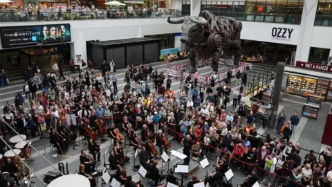PA Media An aerial shot of an orchestra of seated players, who are set up on the concourse of Birmingham New Street station, in the shadow of the huge, mechanical Ozzy The Bull.