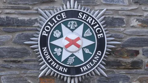 PSNI badge - red cross in white star with green circle and symbols and Police Service of Northern Ireland written around the badge.