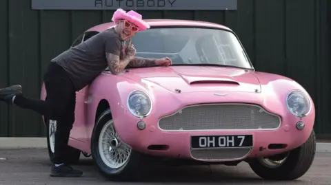 David Pearce-Martin, wearing pink Barbie hat and dark t-shirt and jogging pants draped over the bonnet of a pink 1967 Aston Martin DB6 he has restored. He's wearing pink heart glasses and is smiling to camera.